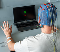 Eine Person sitzt vor einem Computer, hebt die linke Hand und trägt eine Haube auf dem Kopf, aus der elektrische Kabel führen. 